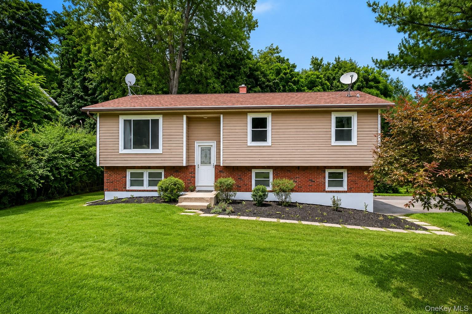 Split-level home featuring a brick and vinyl exterior with multiple windows, a centered entrance with concrete steps, and a professionally landscaped front yard with mature trees, manicured lawn, and established plantings along the foundation