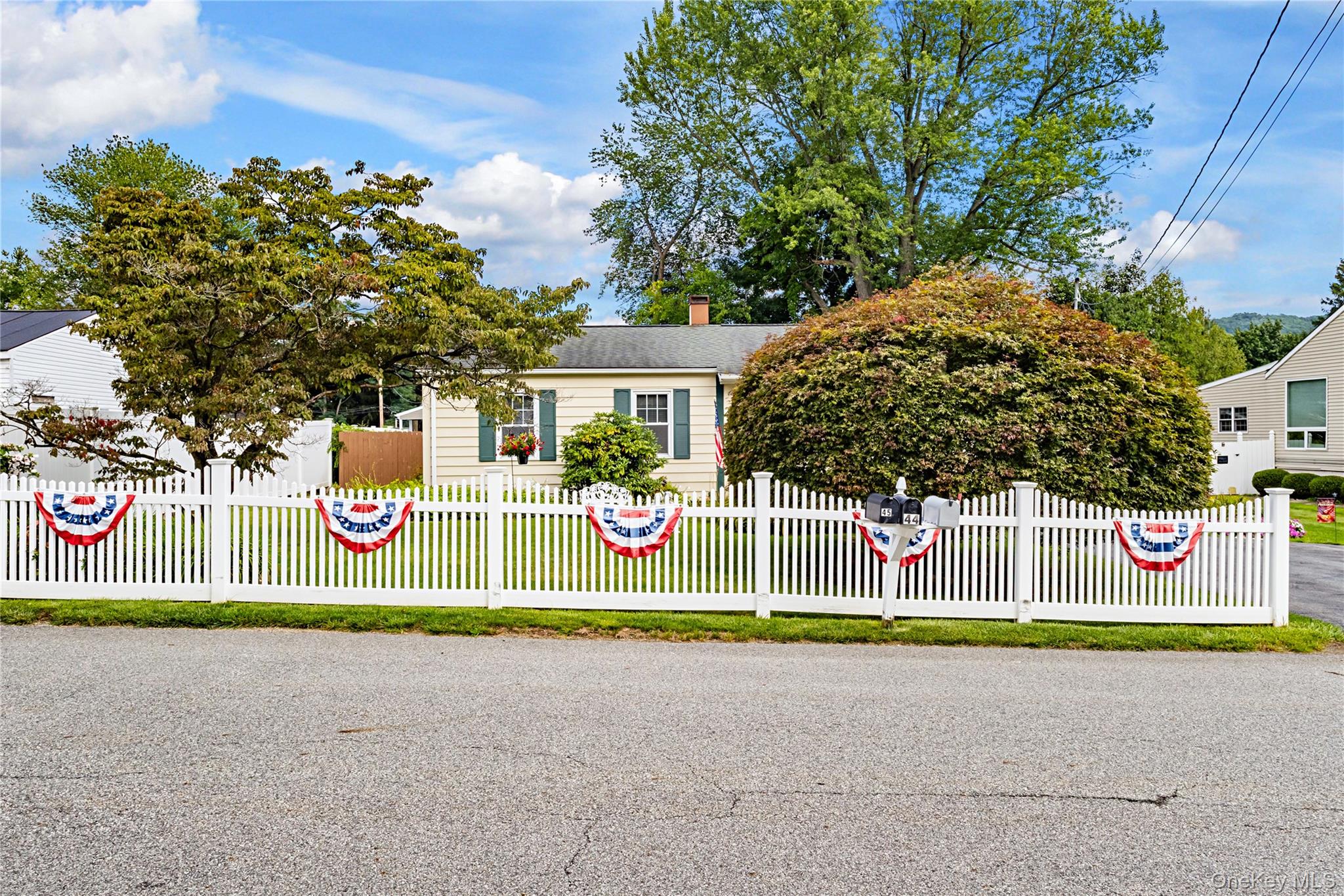 White picket fence enclosing the front yard, featuring a mailbox, mature landscaping, and a pale yellow exterior with green shutters