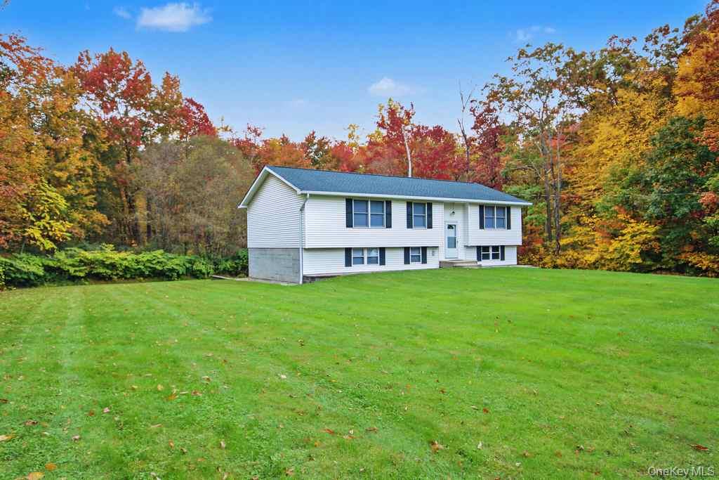 Split-level exterior featuring white siding, dark shutters, a shingled roof, and an expansive lawn set against a wooded backdrop